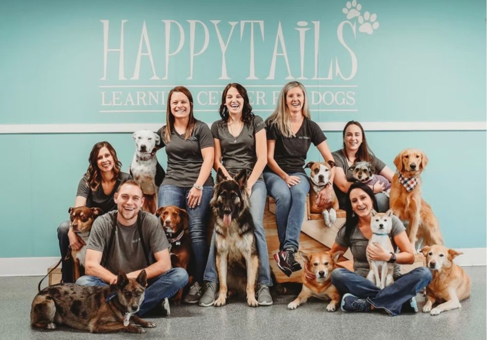 Happy Tails dog training team posing with dogs in front of teal branded wall sign.