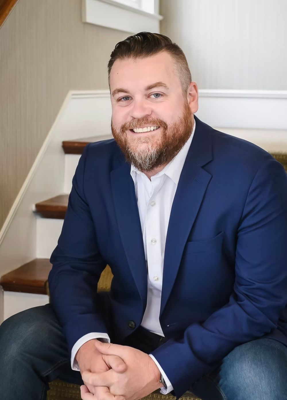 Smiling bearded man in navy blue blazer sitting on staircase steps in professional headshot photo.