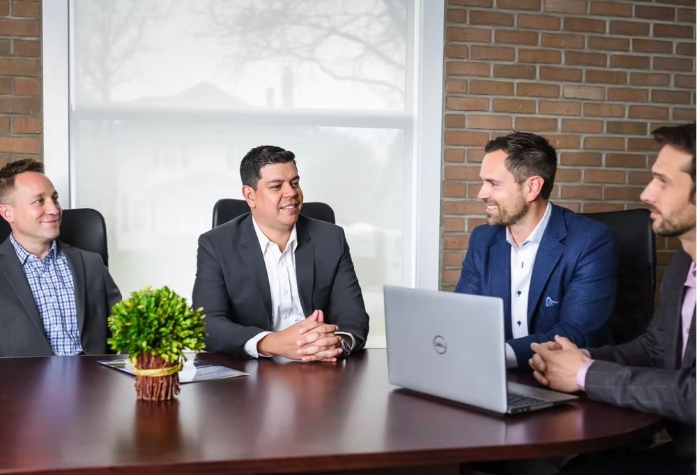 Four businessmen in suits having meeting at conference table with laptop in modern office.