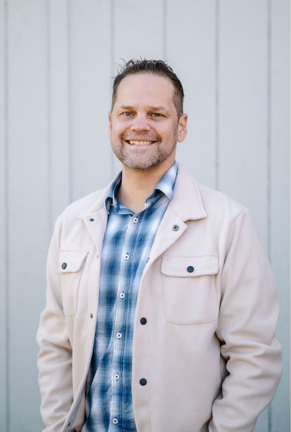 Smiling middle-aged man in beige jacket and plaid shirt posing outdoors against gray wall.