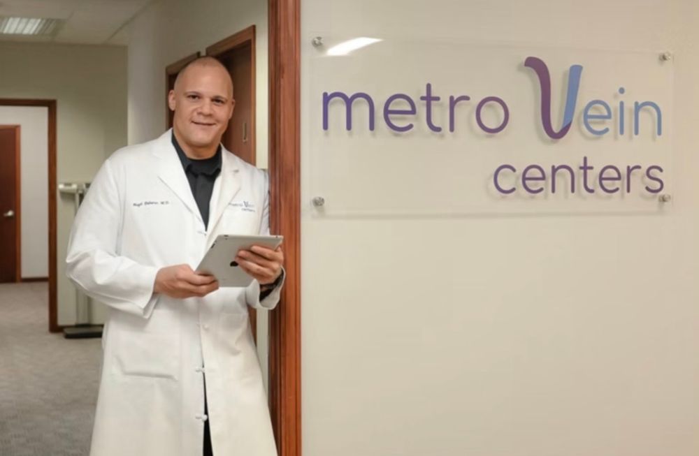 Doctor in white coat holding tablet standing by Metro Vein Centers sign in medical office.