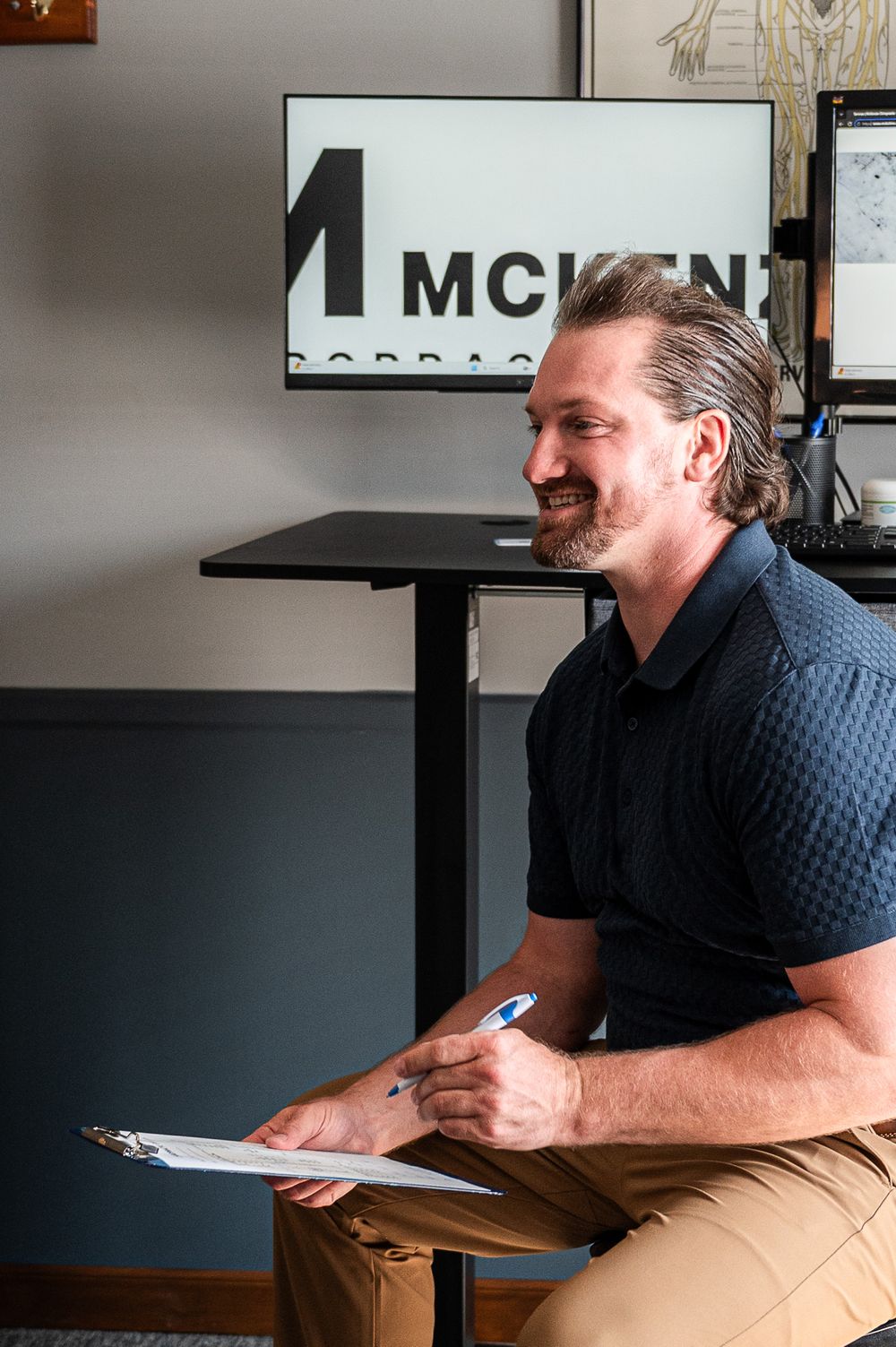 Male chiropractor smiling while holding clipboard seated at standing desk with McKenzie branding sign.
