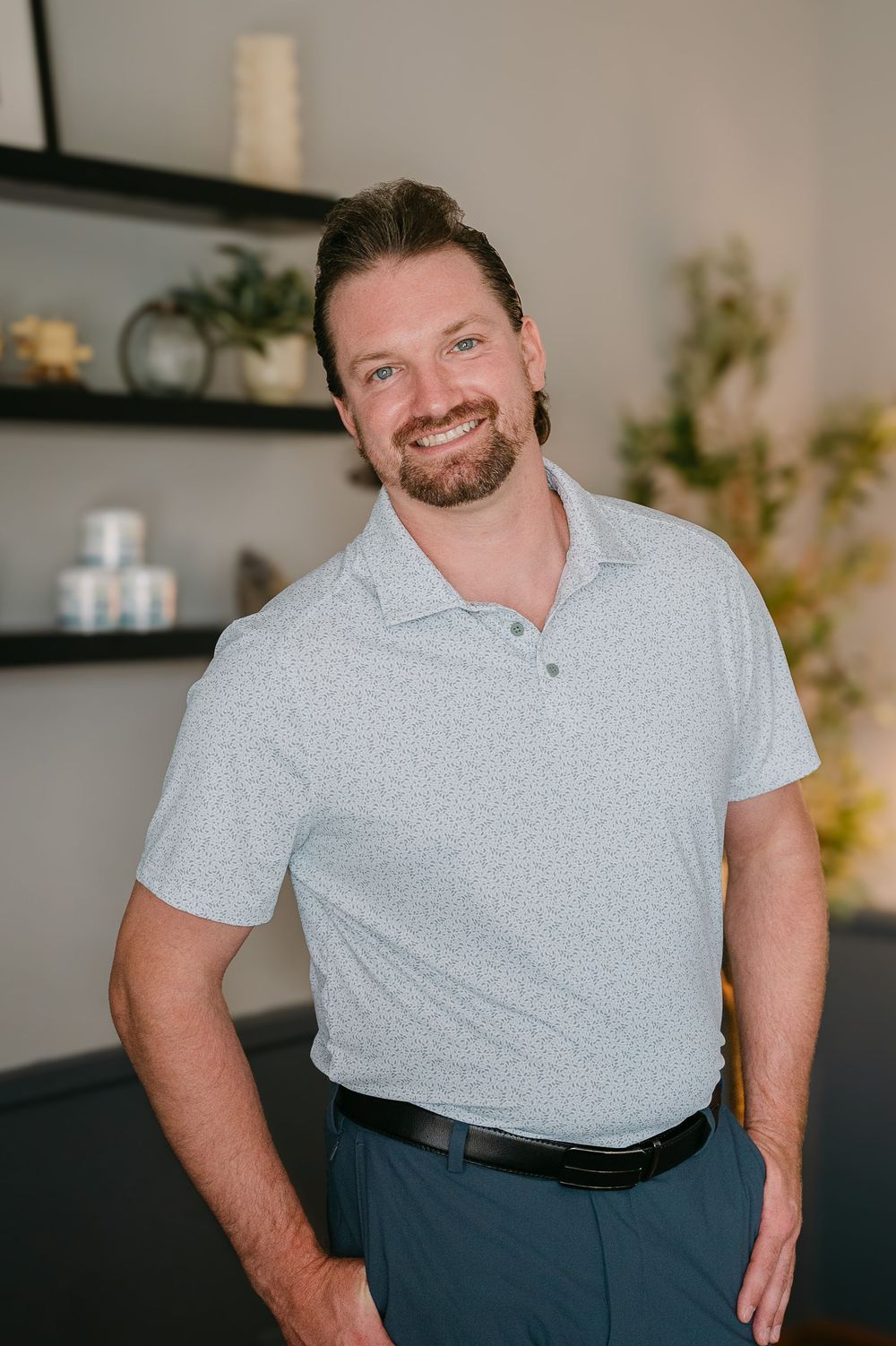Smiling bearded man in light blue polo shirt standing in modern wellness office interior.