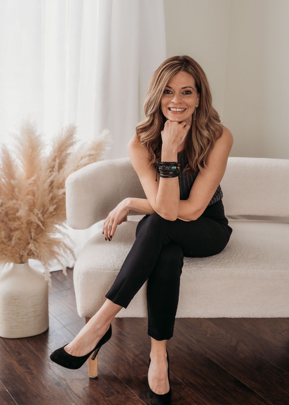 Stylish woman in black outfit sitting on white sofa with pampas grass decor.