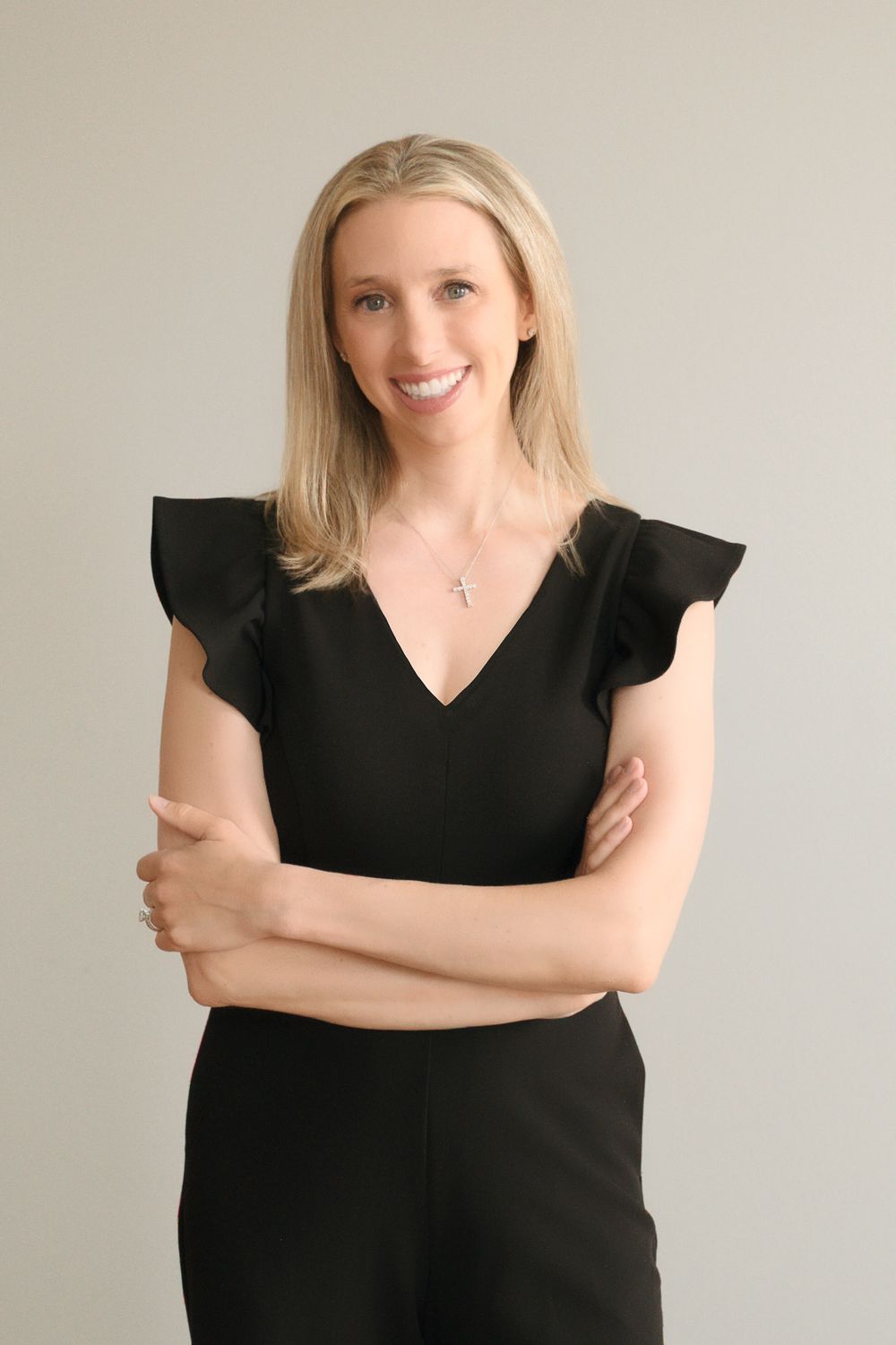 Professional blonde woman in black ruffled dress smiling with arms crossed against gray background.