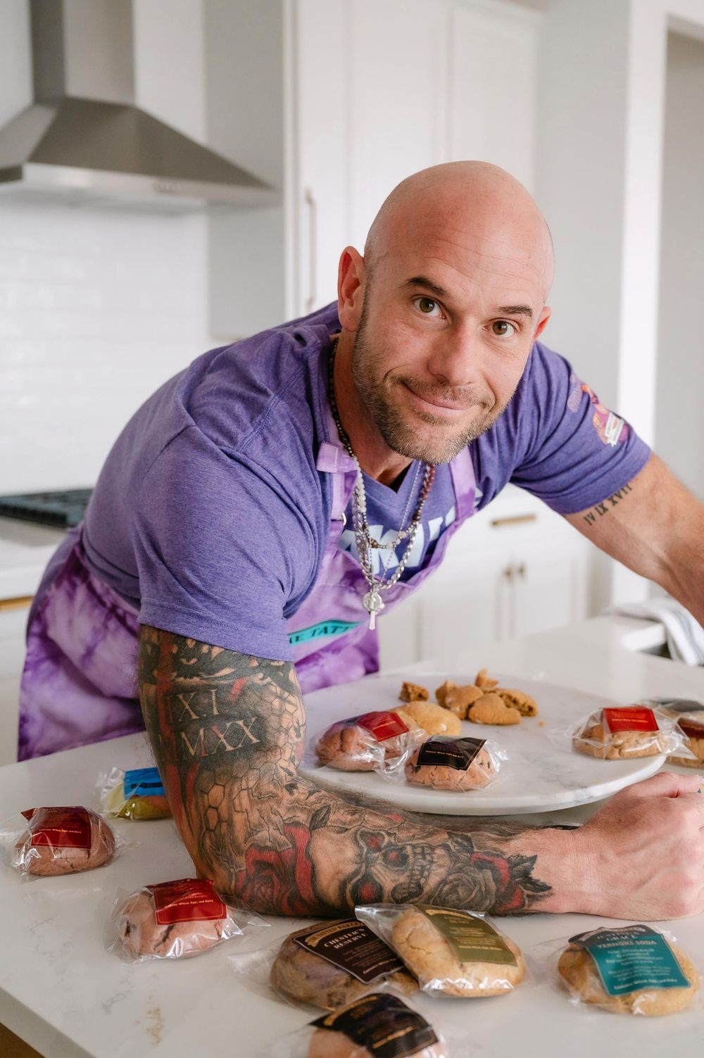 Tattooed bald man in purple apron displaying cookies on kitchen counter.