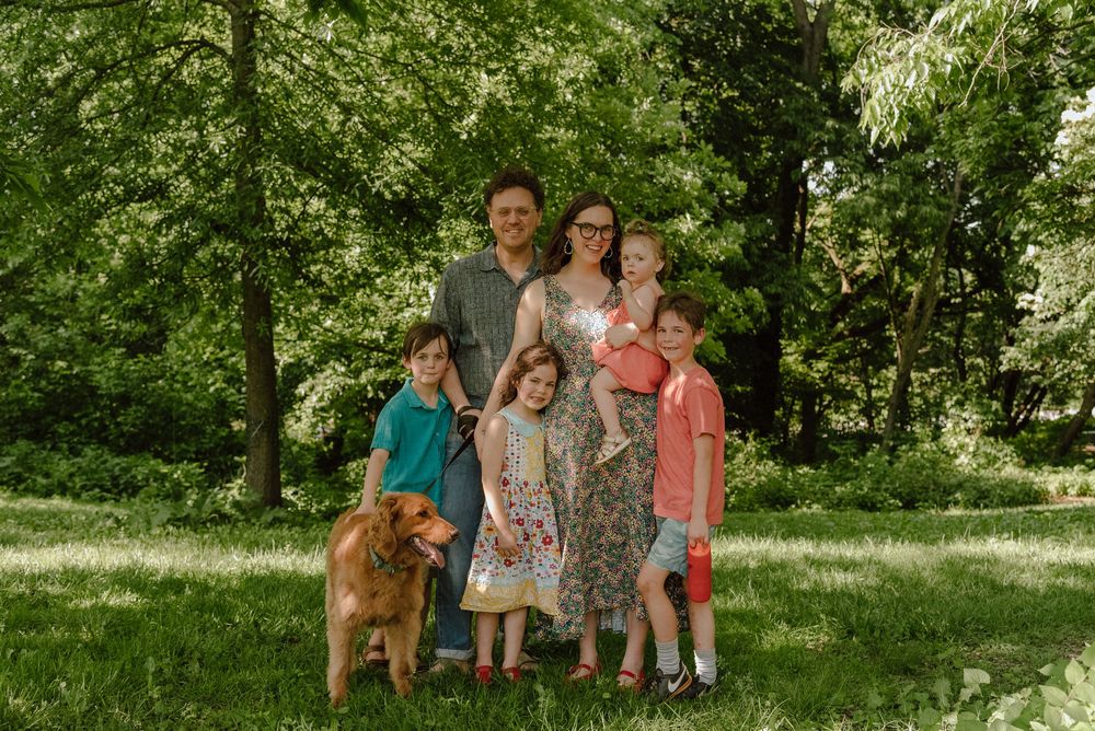 Family of five with a golden dog posing outdoors on green grass surrounded by lush trees on a sunny day.