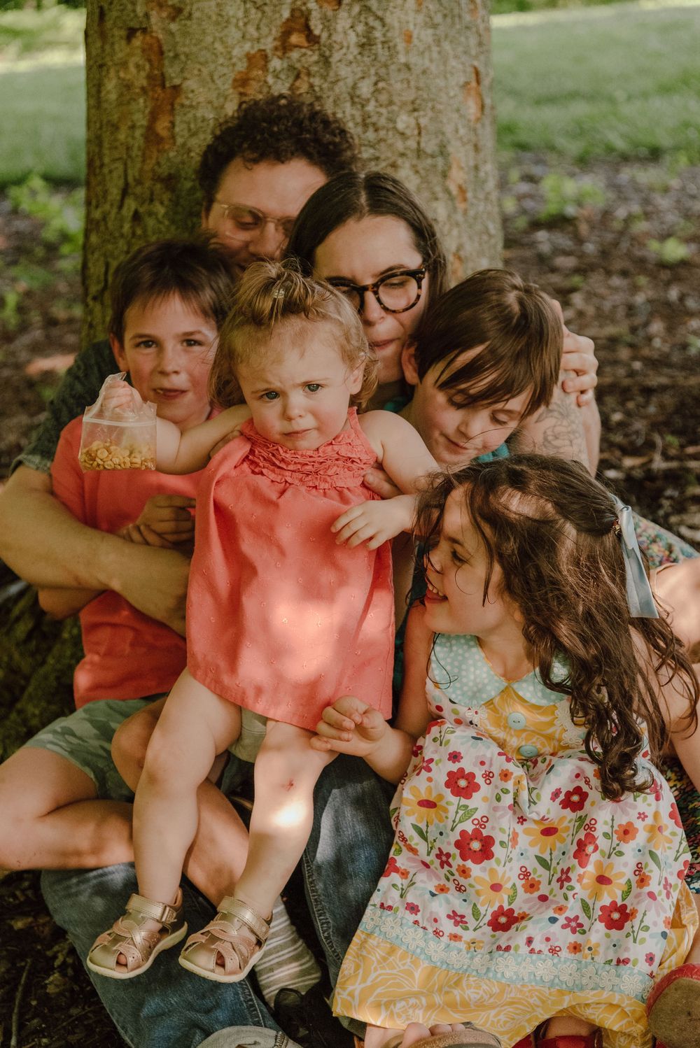 A joyful family of six cuddles together against a tree outdoors, with children and parents sharing warm embraces in nature.