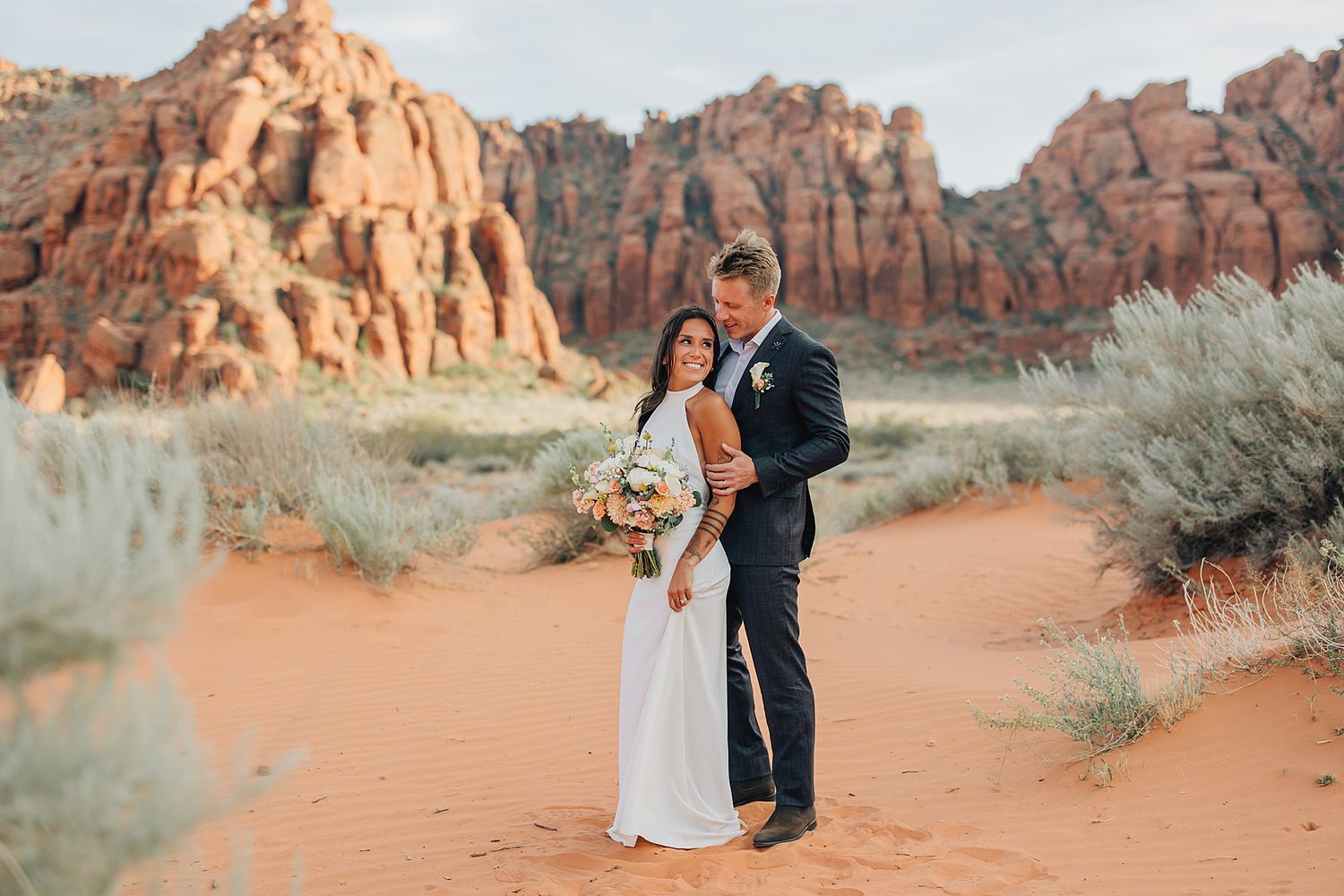 Couple embracing in red sand desert with stunning red rock formations backdrop during outdoor wedding photo session.