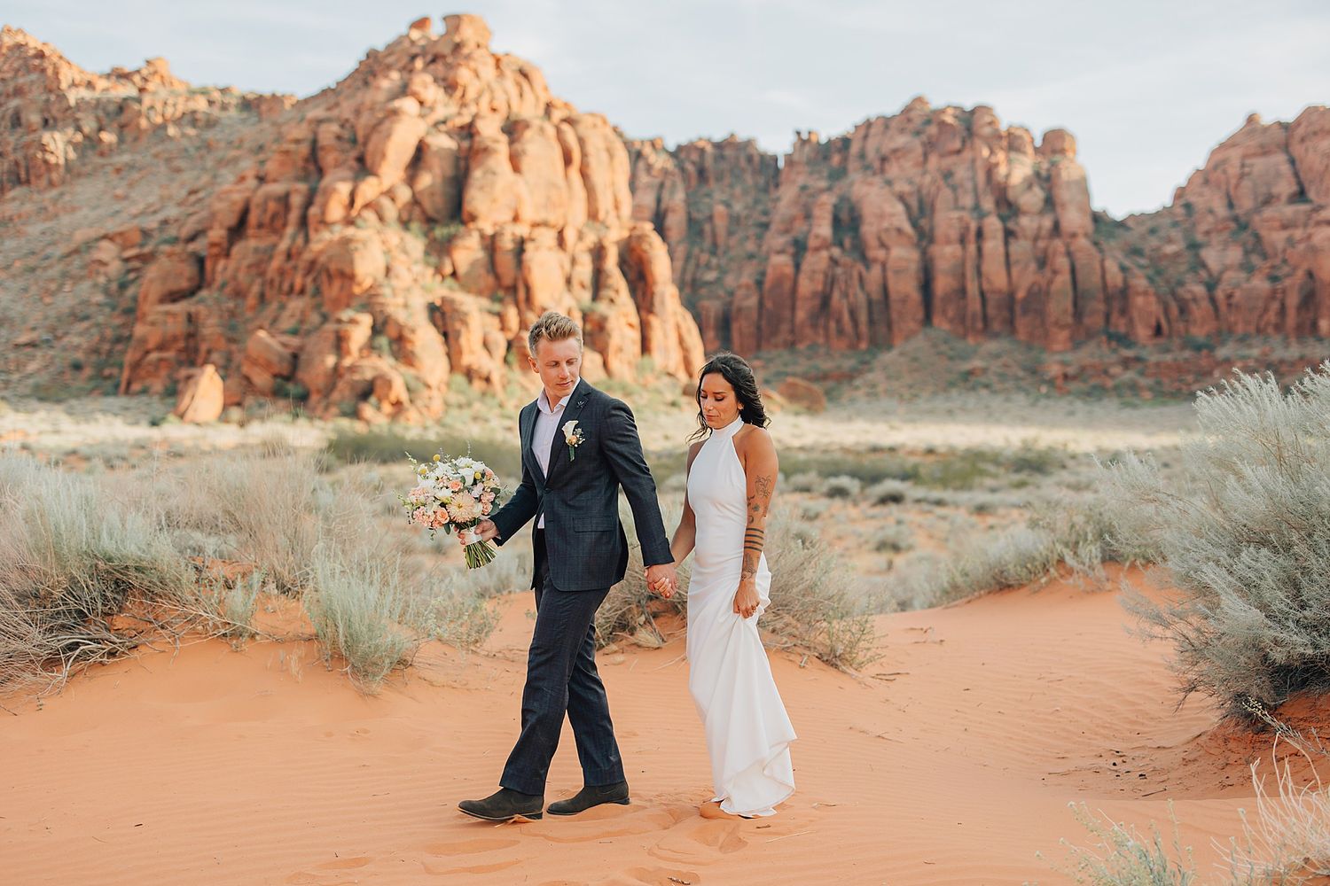 Couple in wedding attire walking on red sand desert with stunning red rock formations in background.