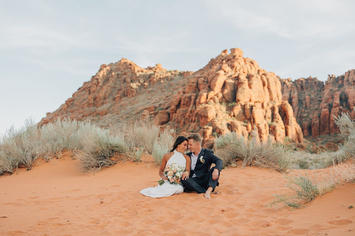 Bride and groom sitting together on red sand dunes with stunning sandstone cliffs backdrop.