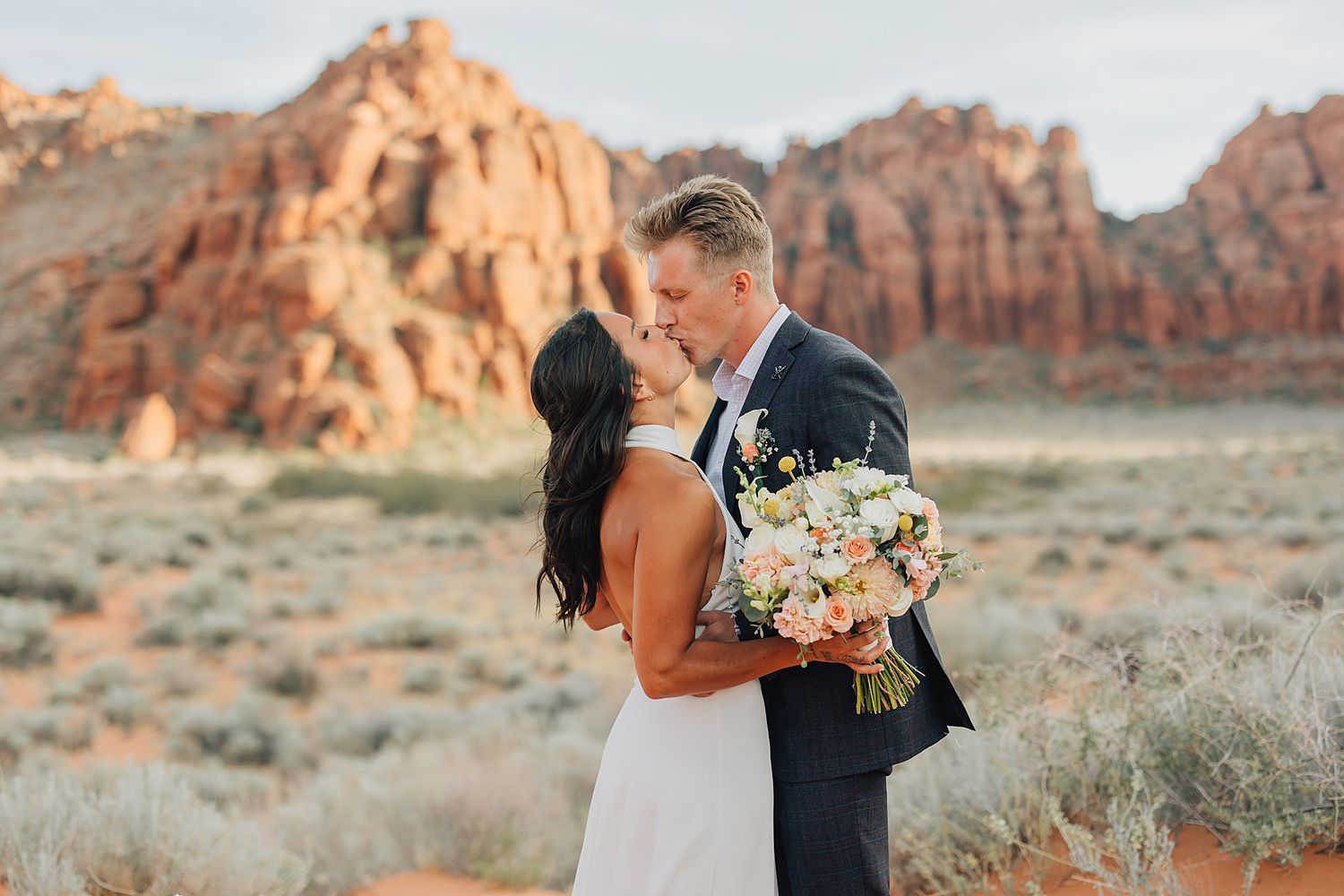 Couple kissing in desert landscape with red rock formations, bride holding floral bouquet, outdoor wedding photography.