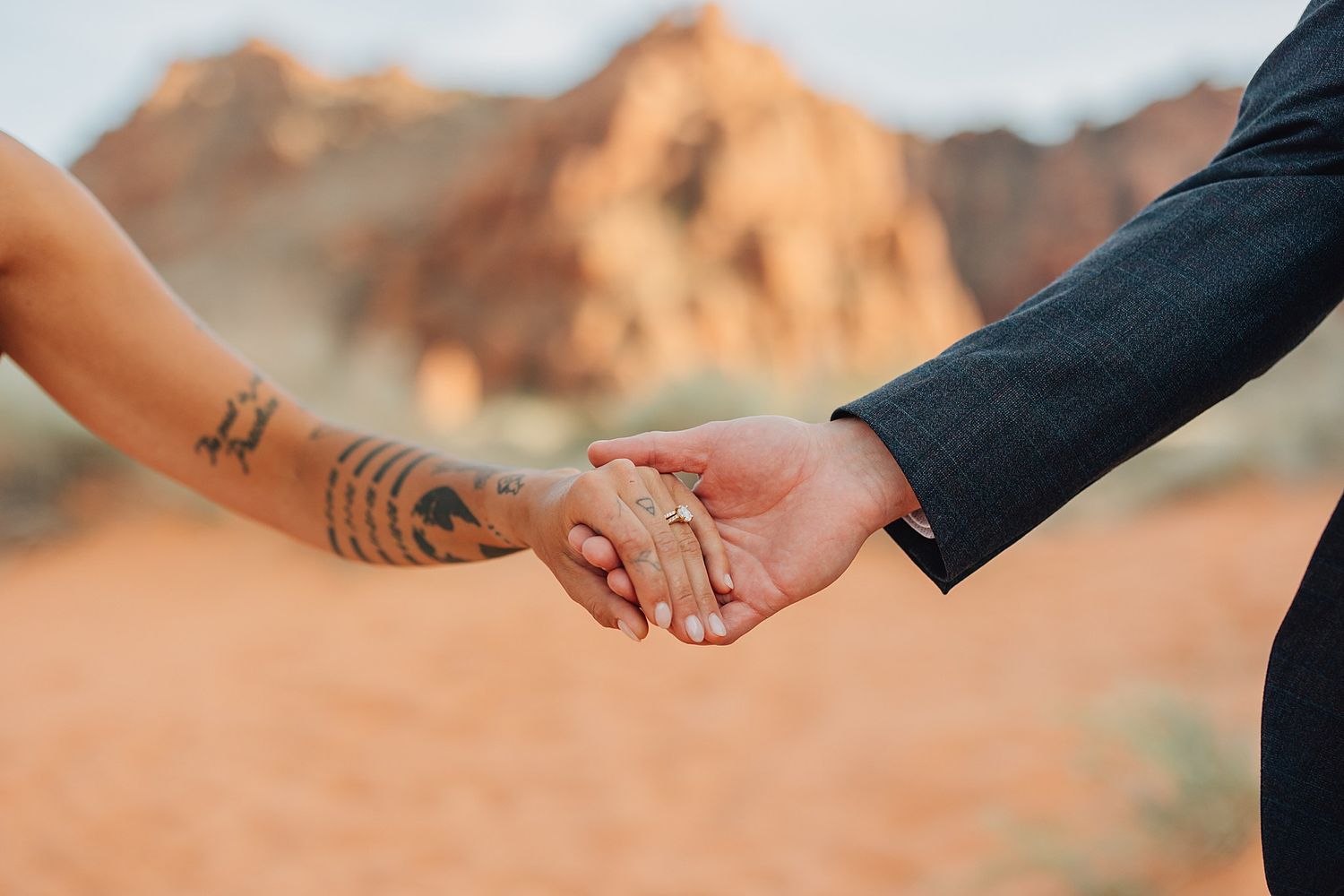 Couple holding hands at desert wedding, bride with tattoos wearing ring, groom in navy suit, red rocks background.