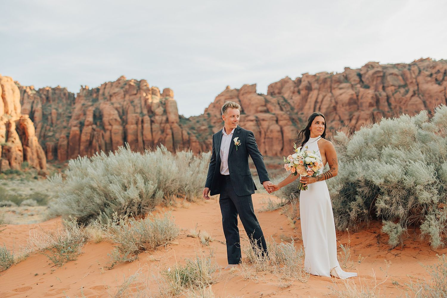 Bride and groom pose in red rock desert landscape with sandstone formations and sagebrush during outdoor wedding.