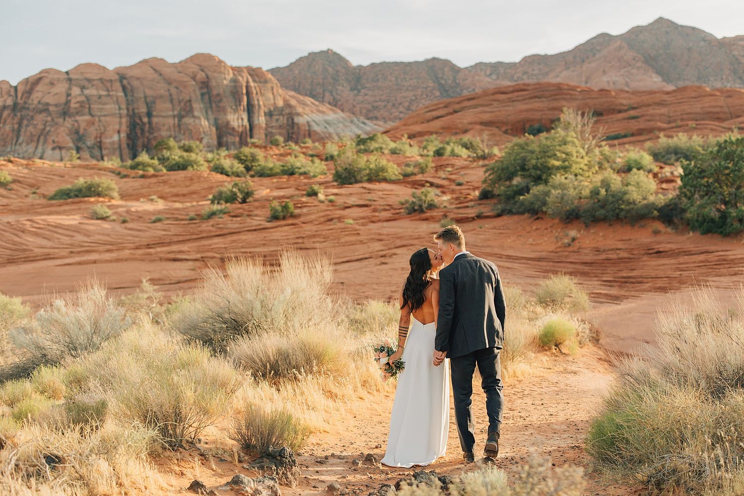 Bride and groom kissing in red rock desert landscape with rustic sandstone mountains in background.