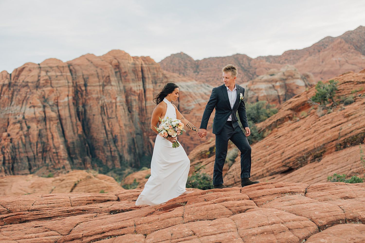 Bride and groom walk hand-in-hand on red rock formations with stunning canyon cliffs backdrop during outdoor desert wedding.