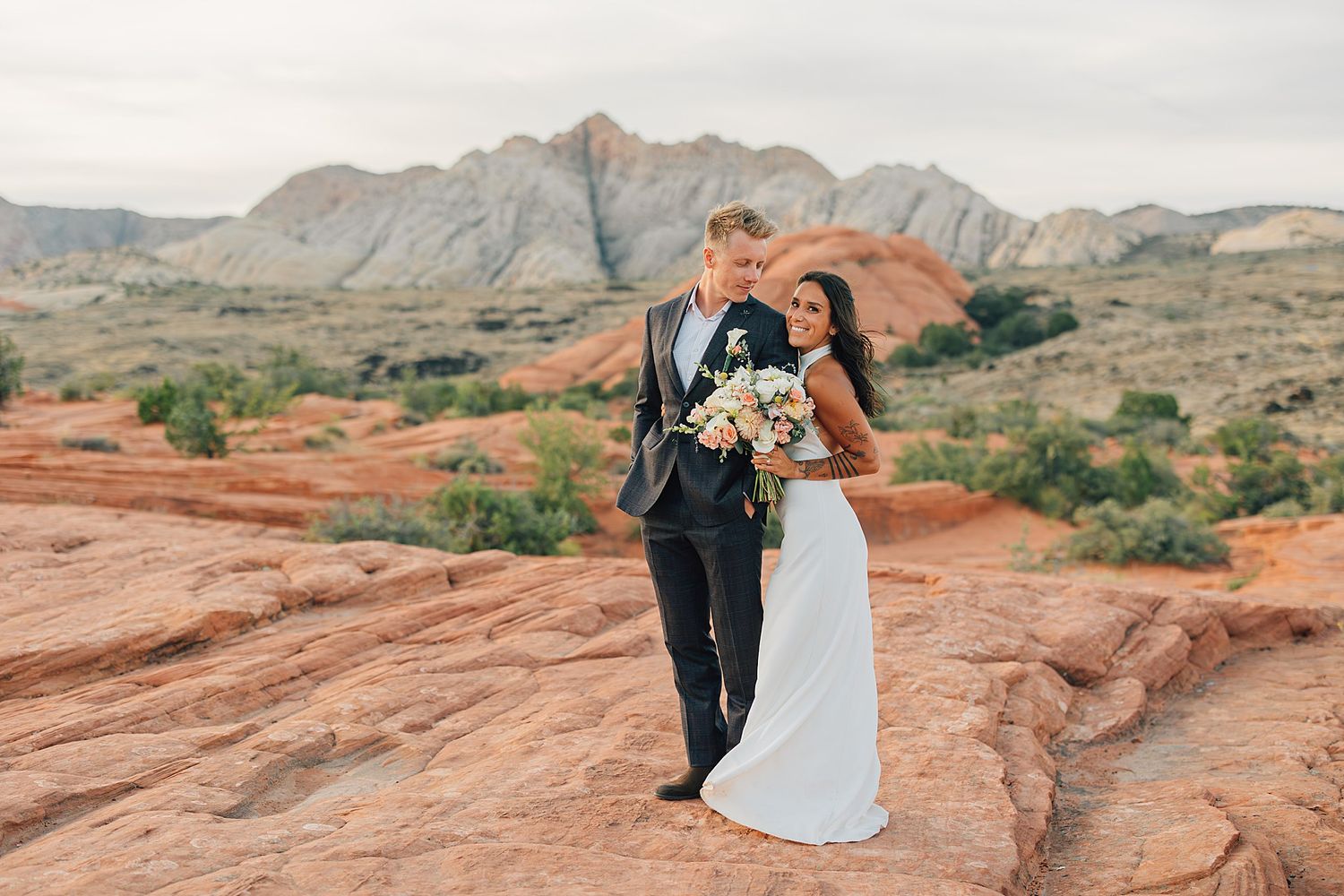 Bride and groom pose on red rock formations with desert mountains backdrop during outdoor wedding ceremony.