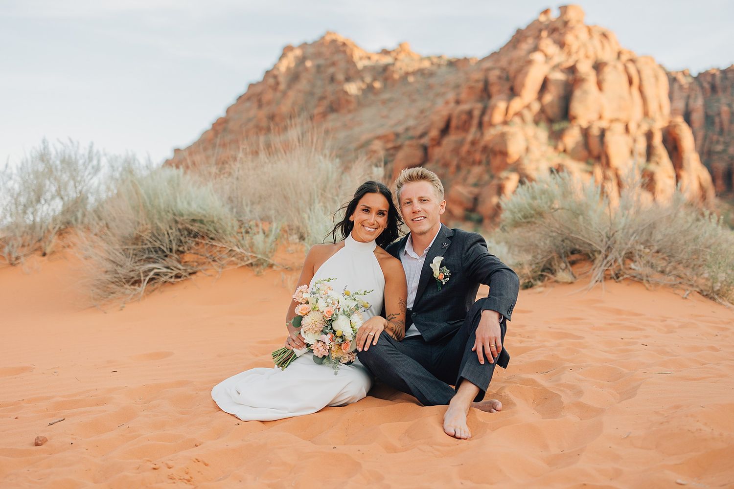 Couple in wedding attire sitting on red sand dunes with rocky desert mountains backdrop during outdoor elopement.