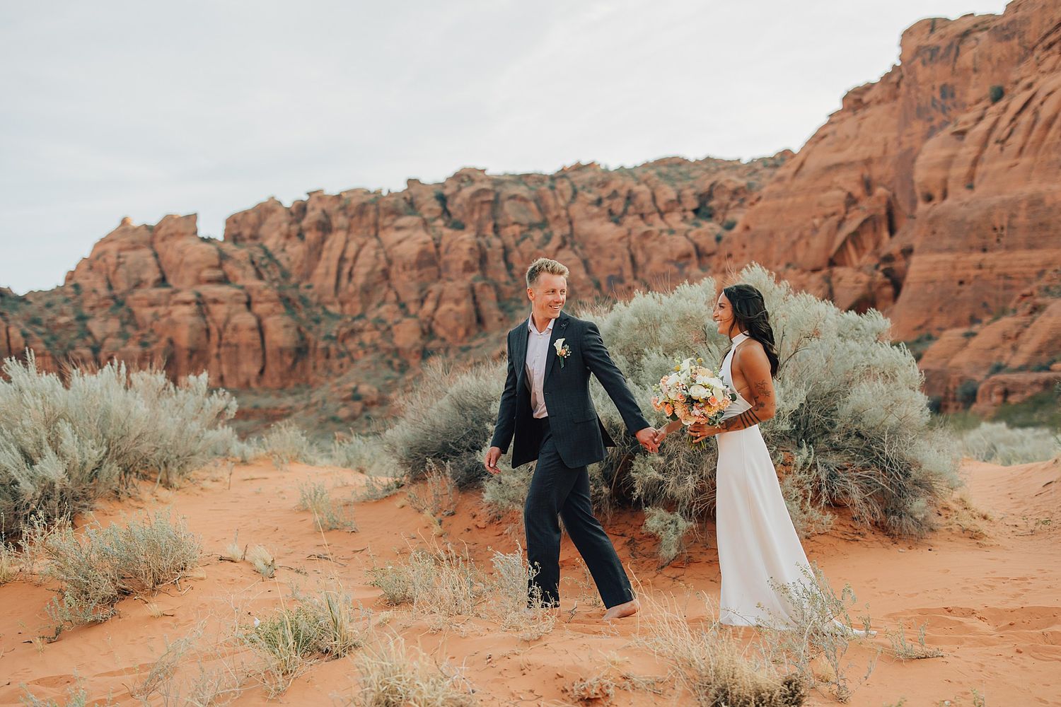 Bride and groom walk hand-in-hand through red rock desert landscape, bride holding colorful bouquet.