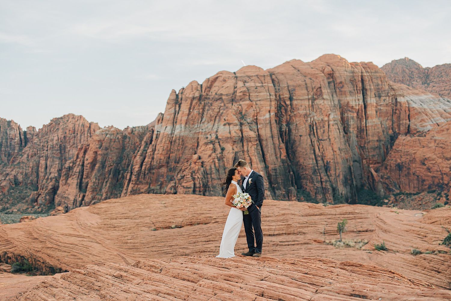 Bride and groom share a romantic kiss on red rock formations with stunning desert canyon backdrop.