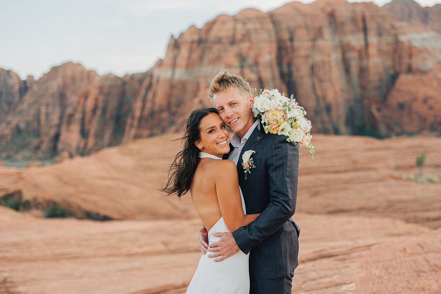 Bride and groom embrace smiling in front of red rock canyon landscape during outdoor desert wedding photo session.
