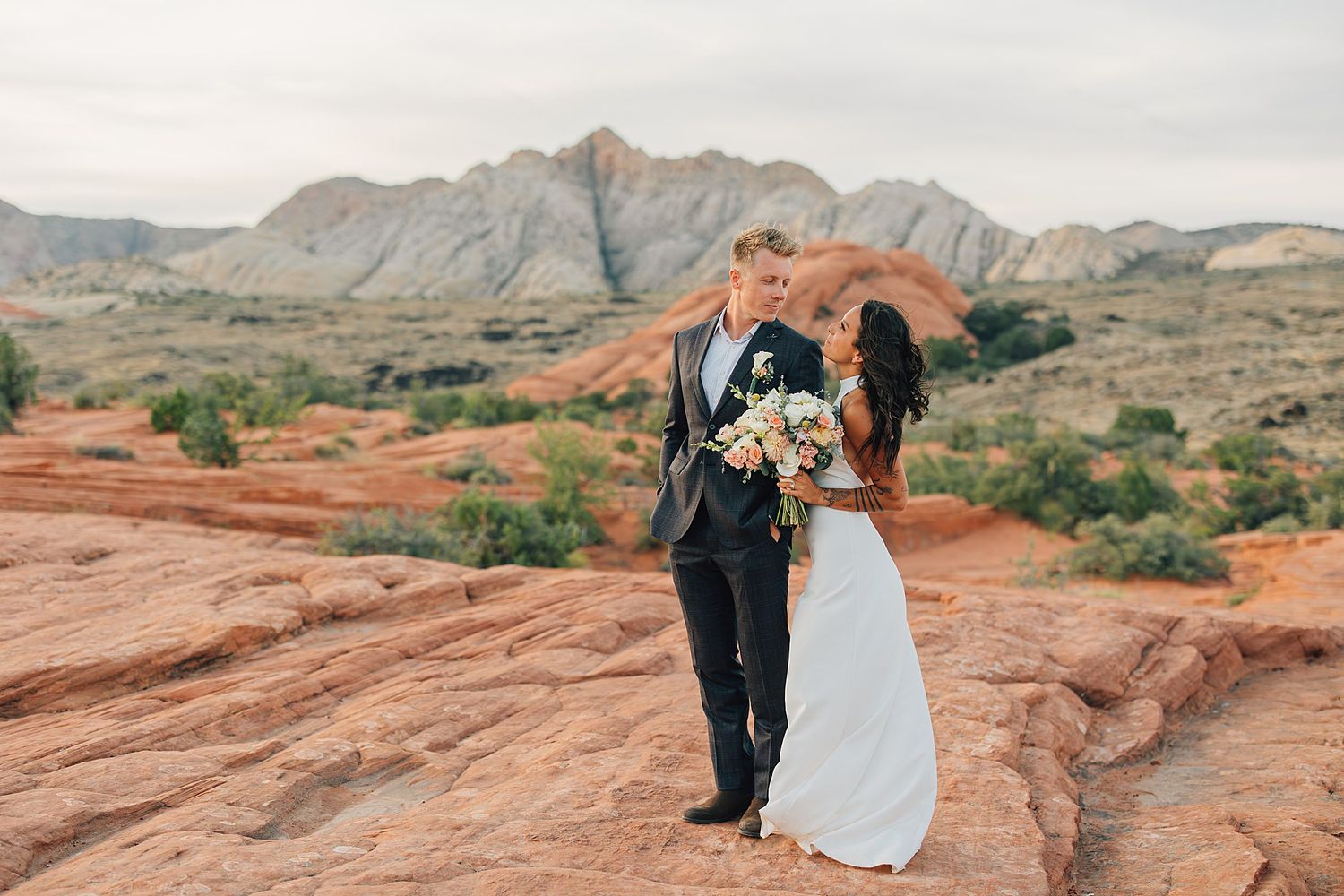 Bride and groom share a romantic moment on red rock formations with stunning desert mountains in the background.