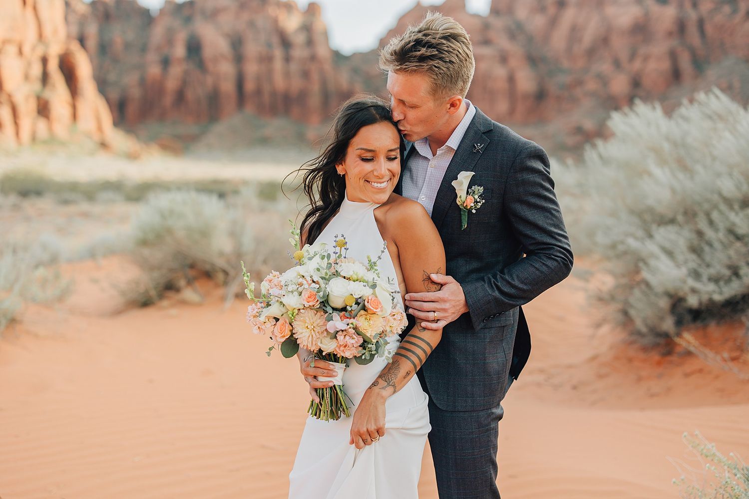 Couple embracing in red rock desert landscape, bride holding pastel bouquet, groom in navy suit kissing her cheek.