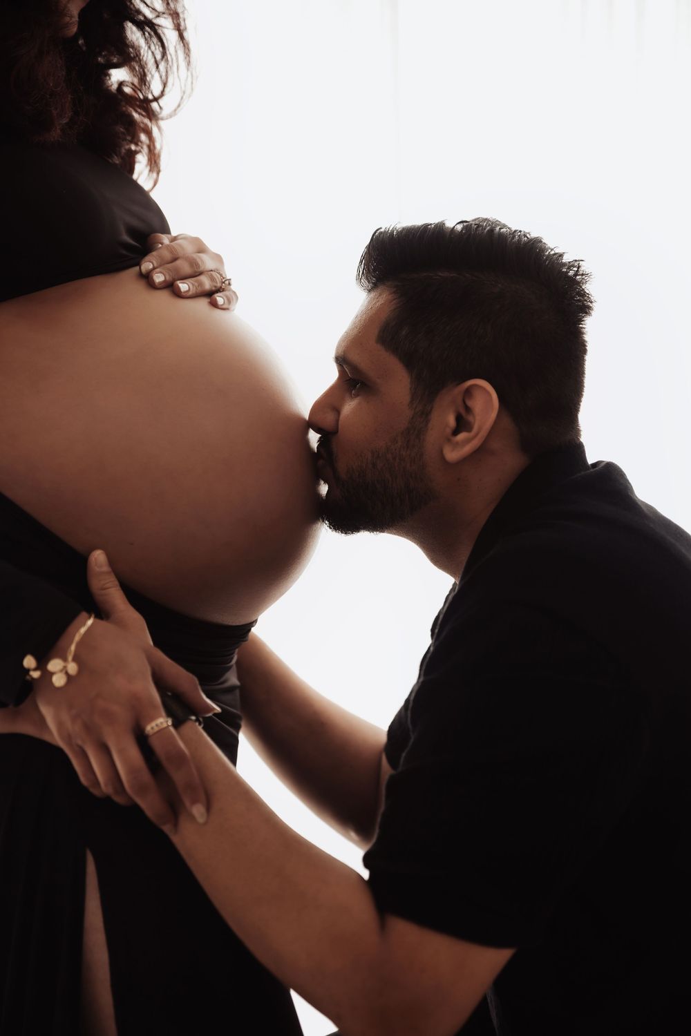 A man tenderly kisses a pregnant woman's baby bump in a beautiful maternity photo shoot with soft backlighting.