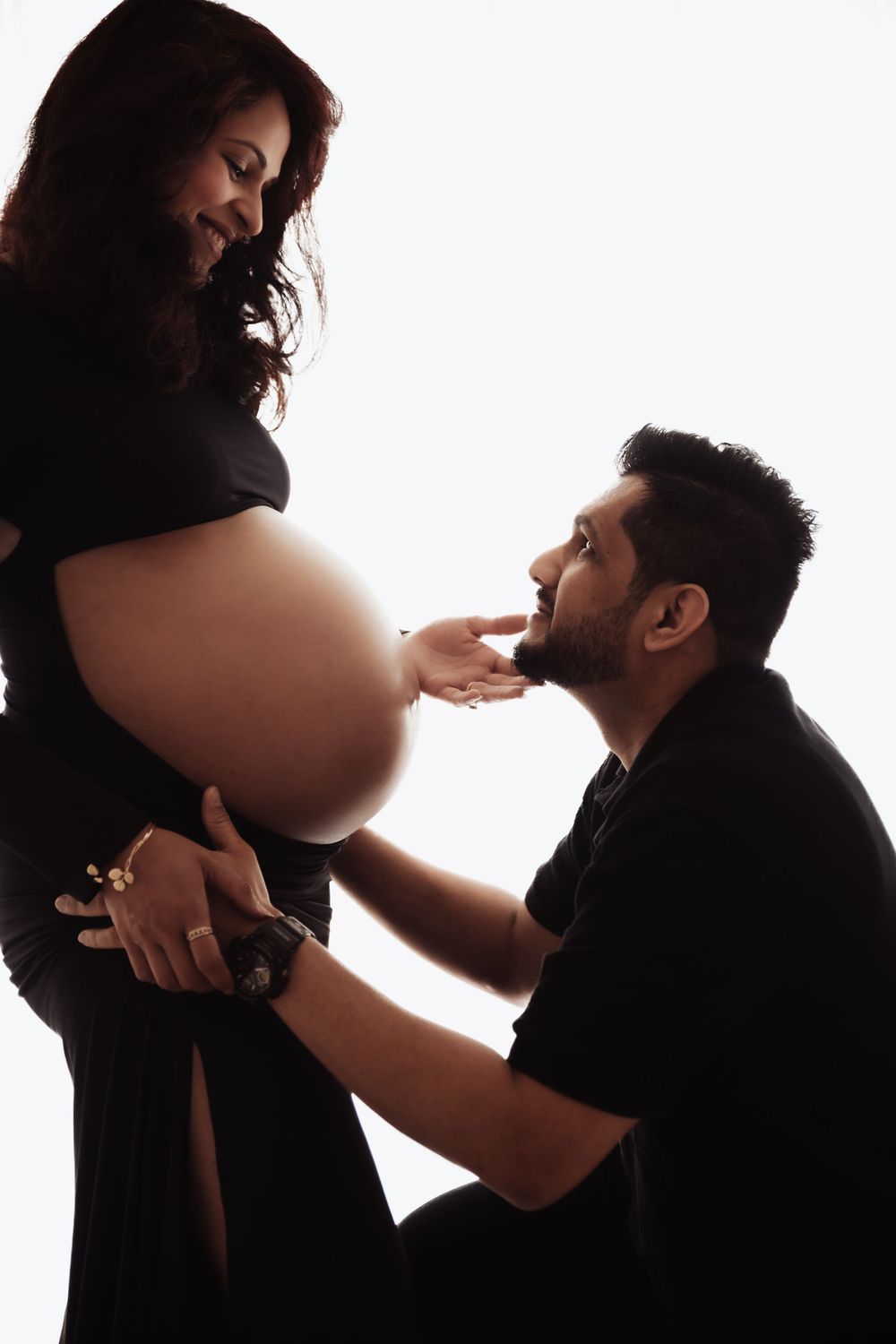 A man kneeling and tenderly touching a pregnant womans bare belly, both dressed in black against a white background.