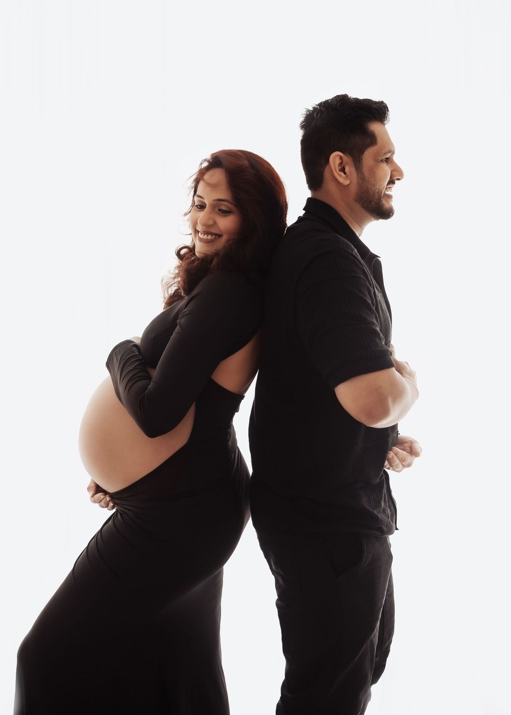 Pregnant woman in black dress poses back-to-back with smiling man in black shirt during maternity photoshoot.