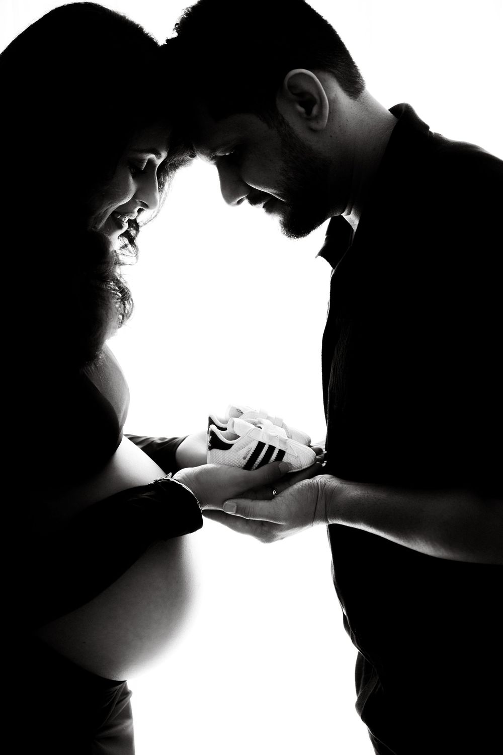 Black and white silhouette of expecting couple holding tiny baby shoes together in bright studio light.