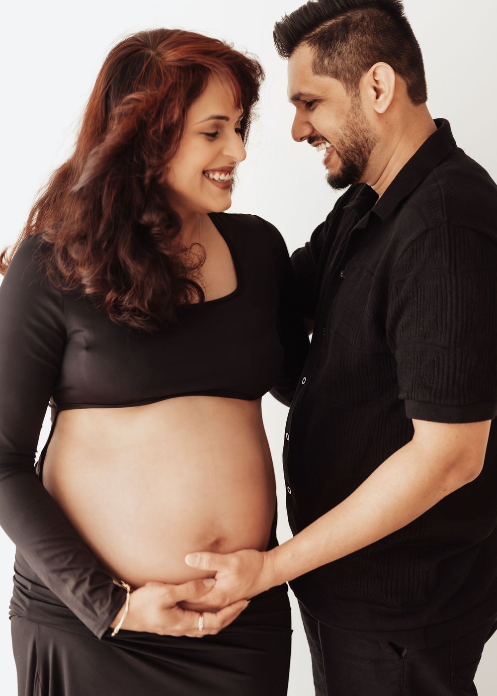 Happy pregnant couple in black outfits holding baby bump together, smiling at each other in a studio setting.