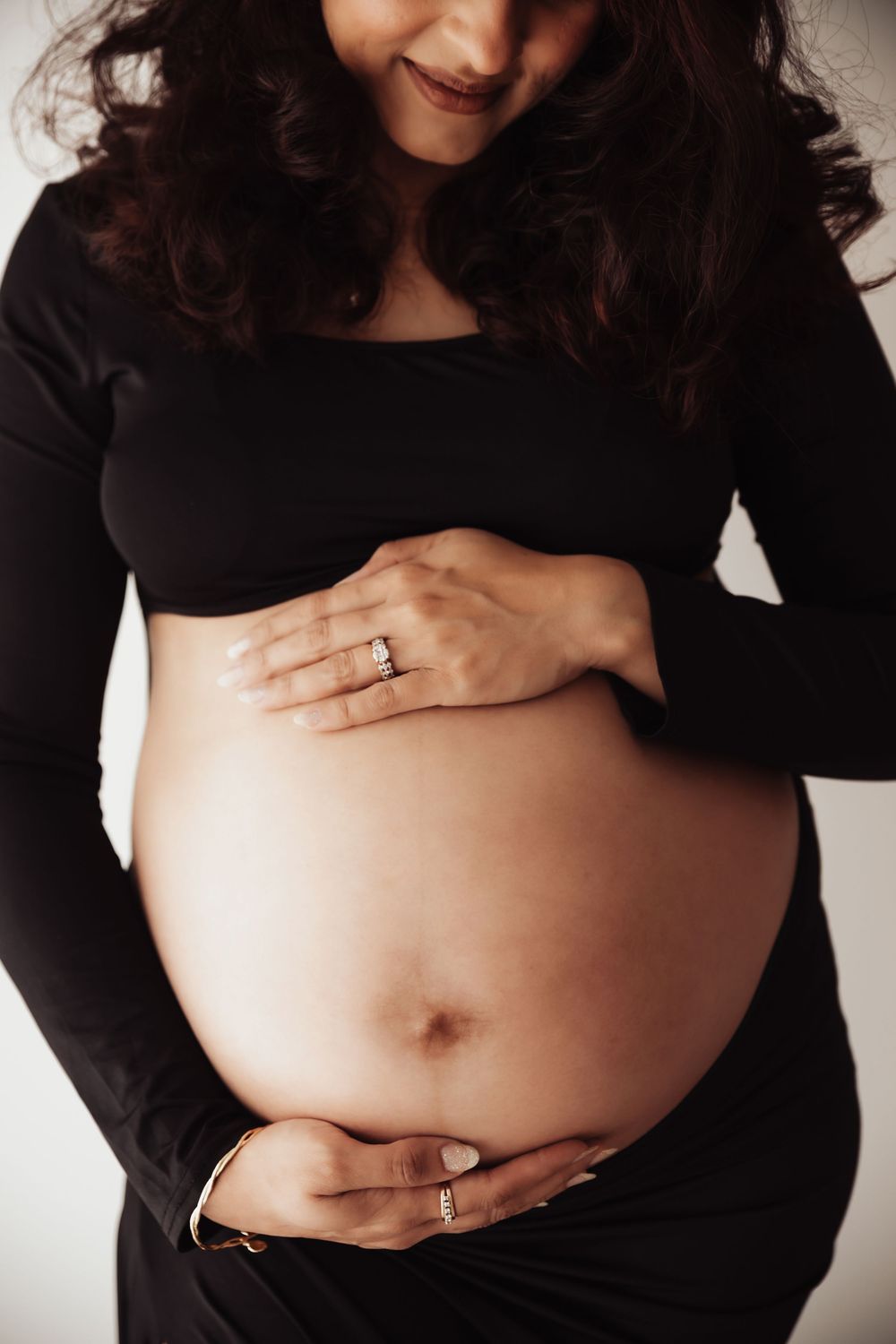 Pregnant woman in black outfit cradling her bare baby bump with both hands, wearing a diamond ring and gold bracelet.