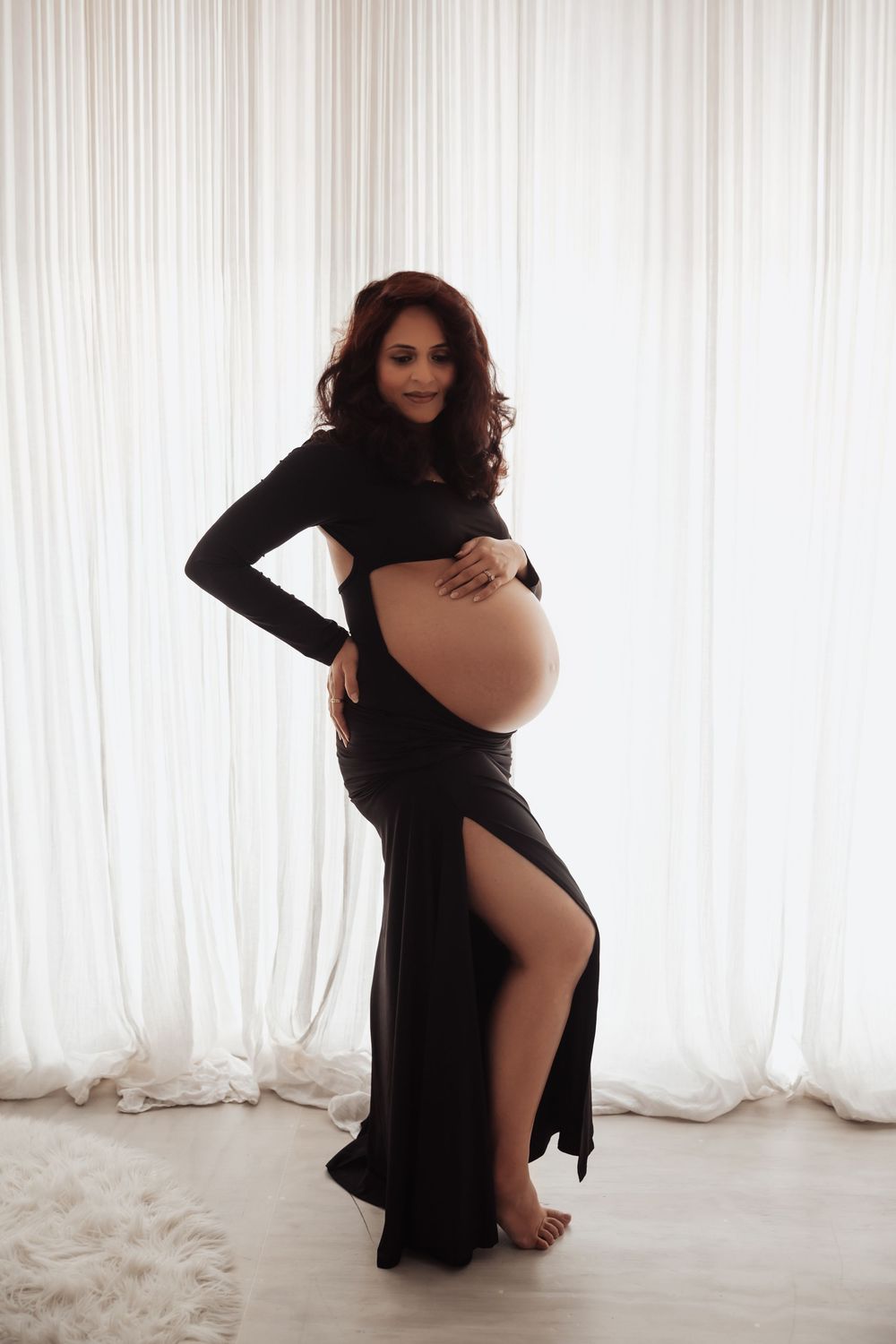 Pregnant woman in black cutout gown cradling bare baby bump, posing in bright studio with white curtain backdrop.