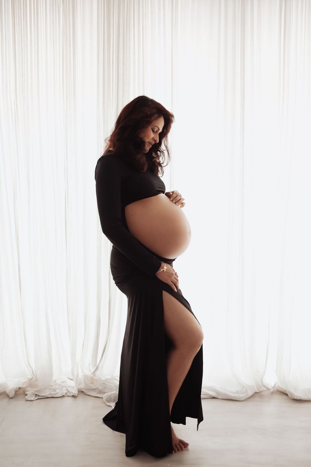 Side profile of pregnant woman in elegant black dress holding baby bump, silhouetted against white curtains.