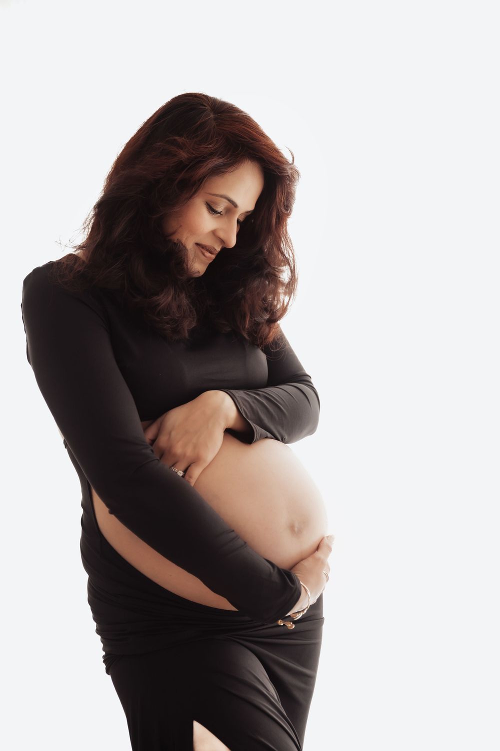 Pregnant woman in black dress cradling her baby bump, smiling softly against a white background.