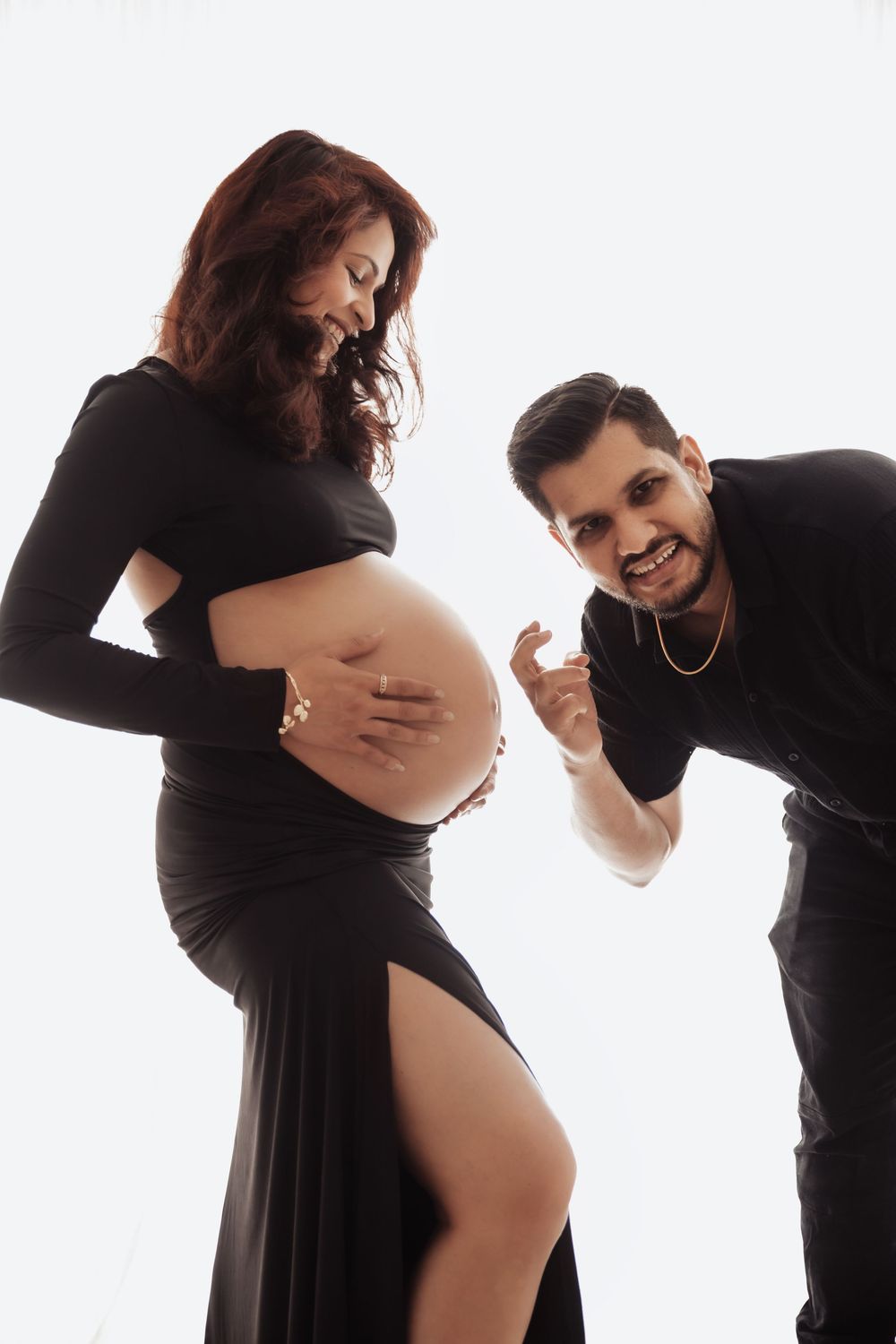 A pregnant woman in a black dress cradles her baby bump while her smiling partner leans in joyfully during a maternity photoshoot.