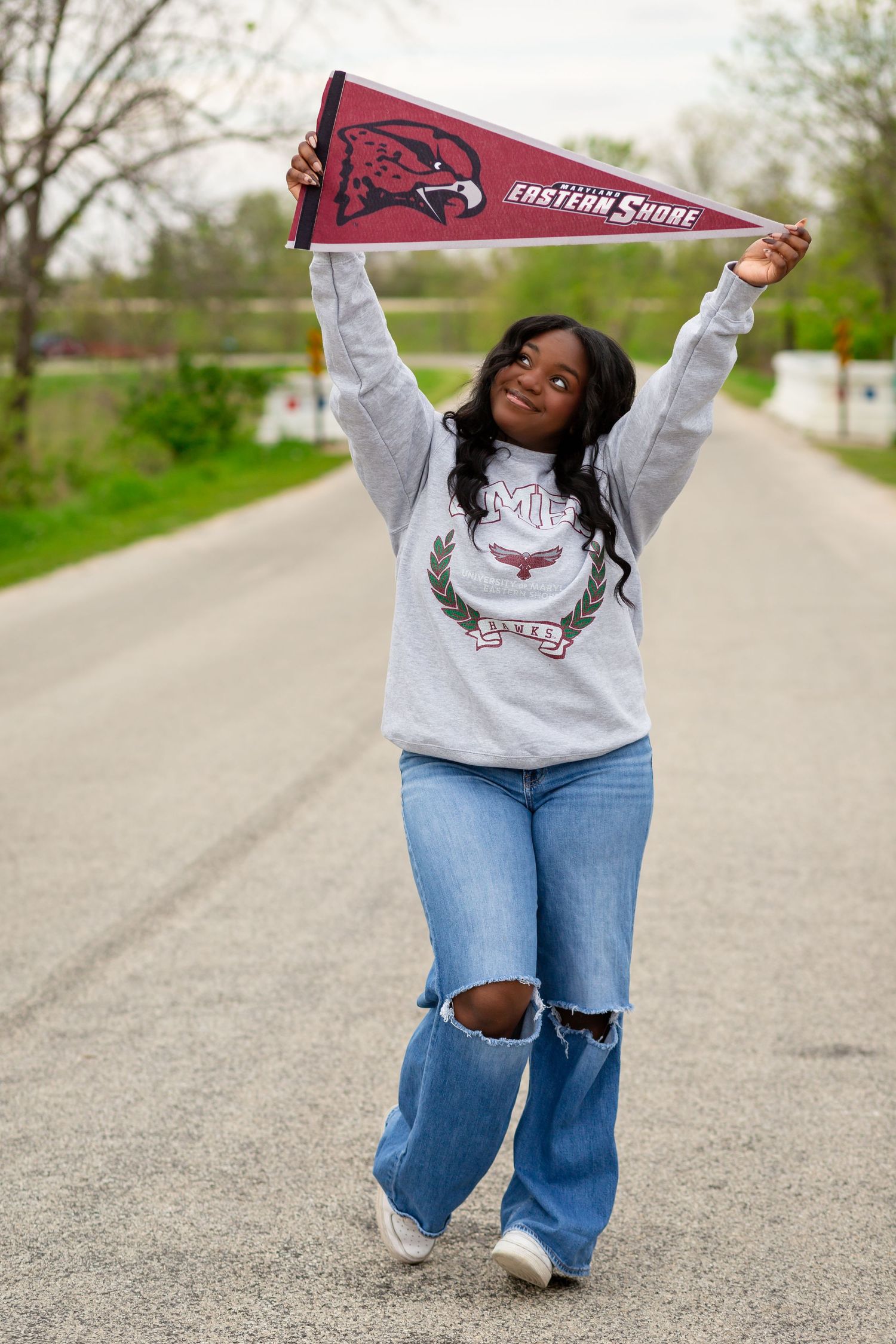 Young woman proudly holding a University of Maryland Eastern Shore pennant overhead while walking on a road in jeans and sweatshirt.