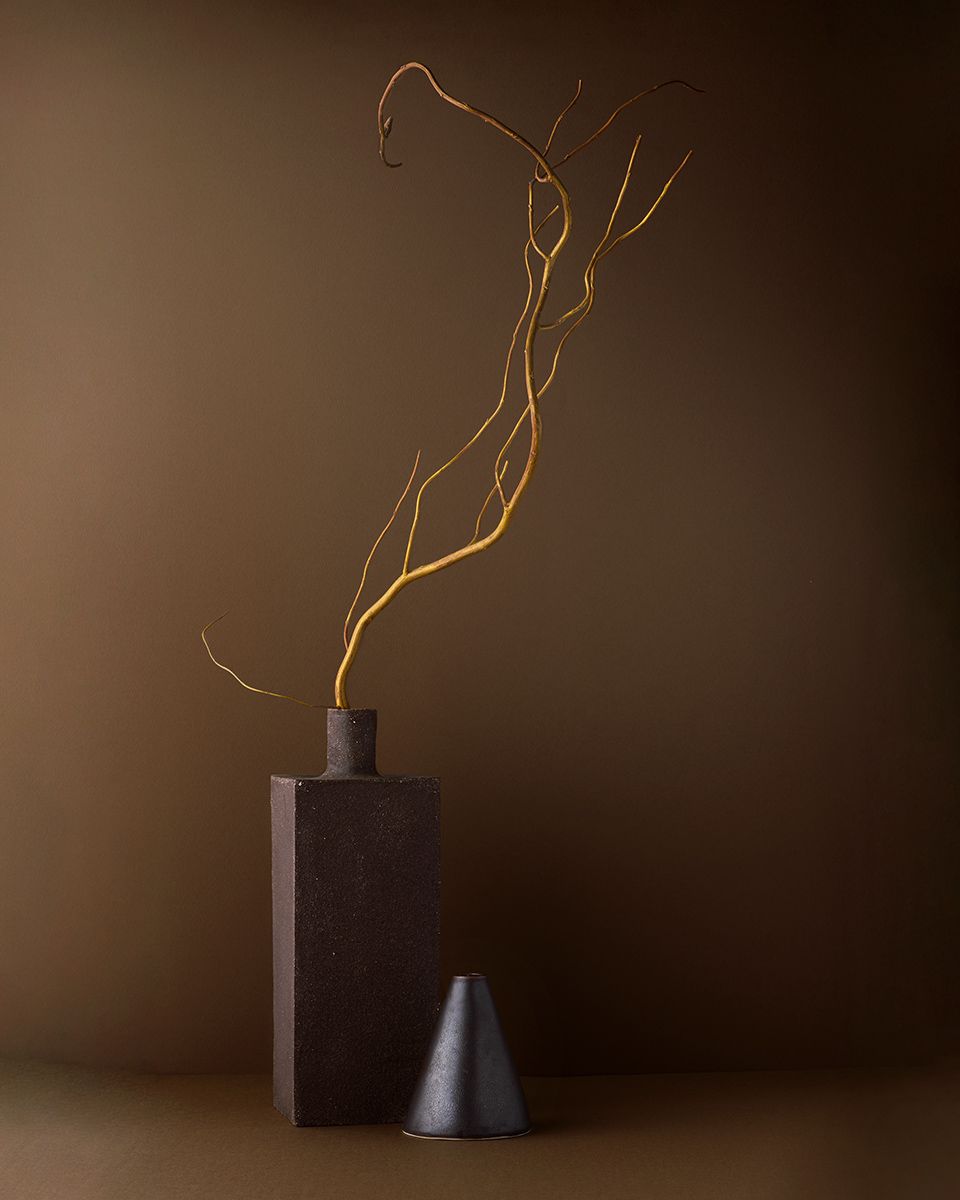 Dark ceramic vase with golden curving branches beside a small conical vessel on a warm brown background.