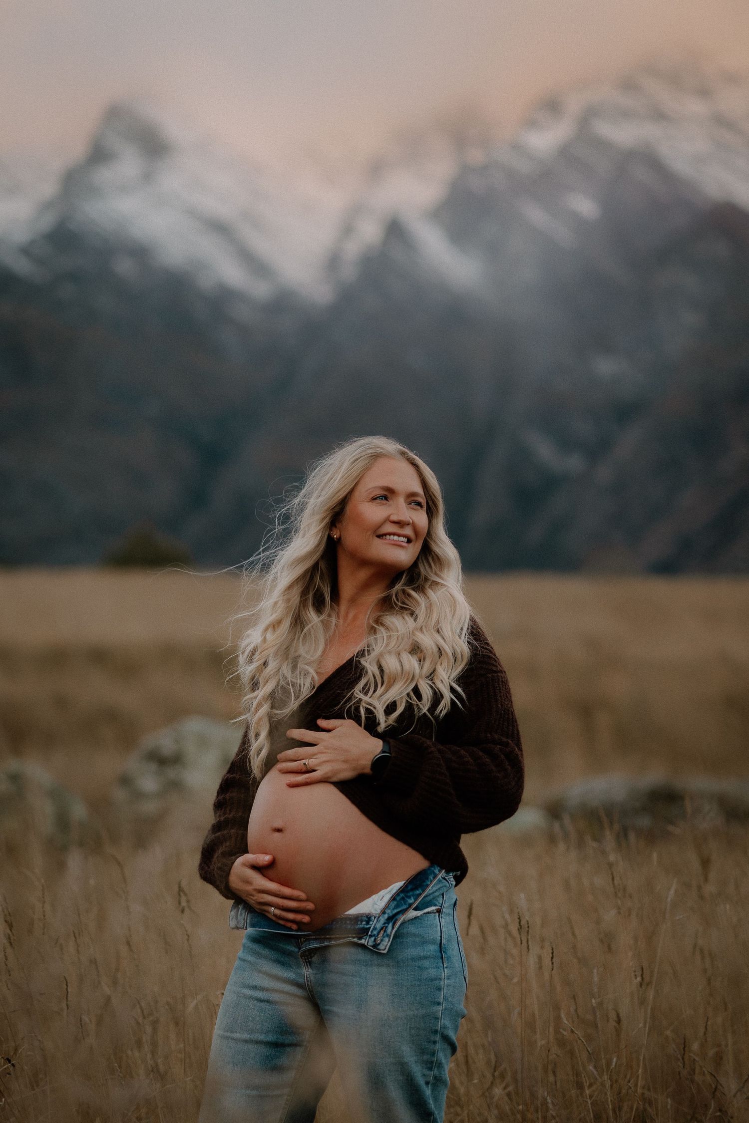 Pregnant woman in brown sweater and jeans cradles baby bump in golden field with snowy mountains at sunset.