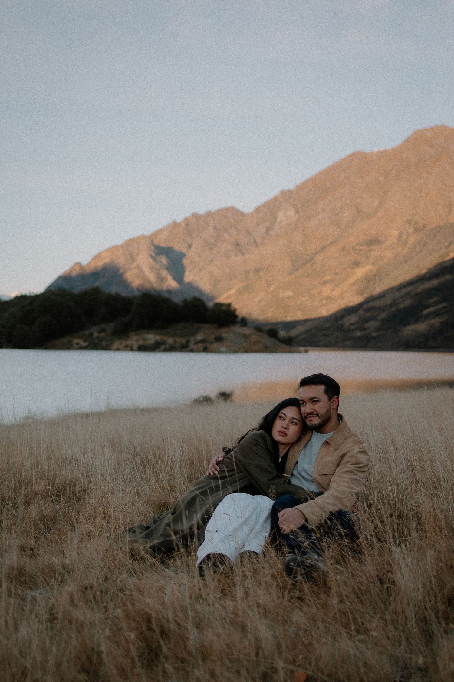 Couple sitting in golden grass by a mountain lake at dusk, embracing romantically with sunlit peaks in background.