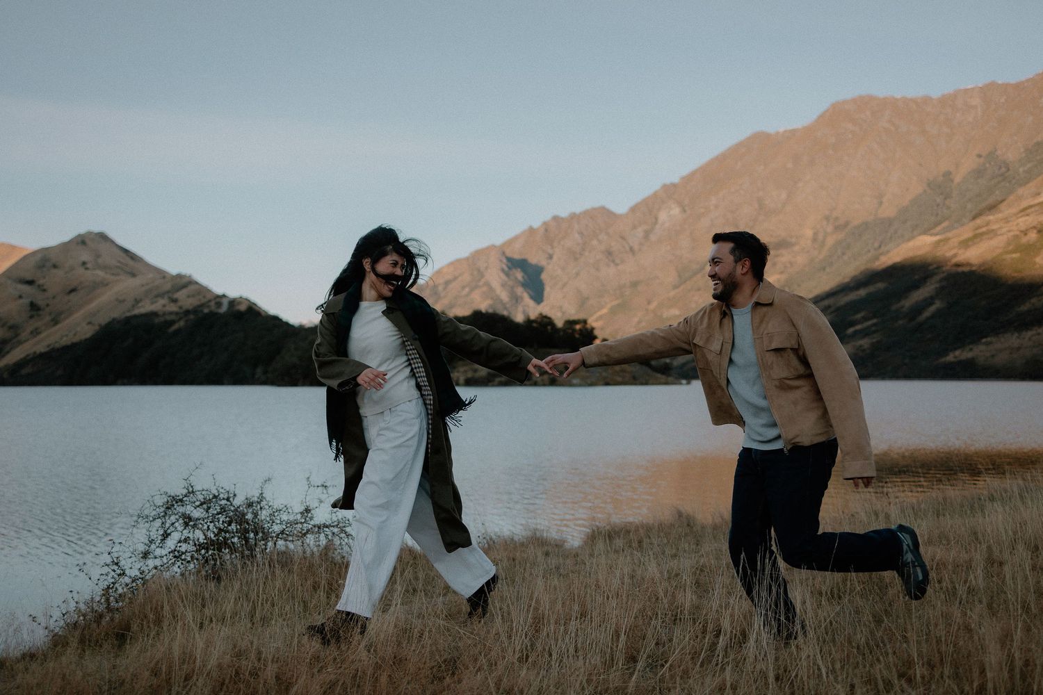 Couple holding hands and running playfully by a mountain lake in golden grass at dusk.