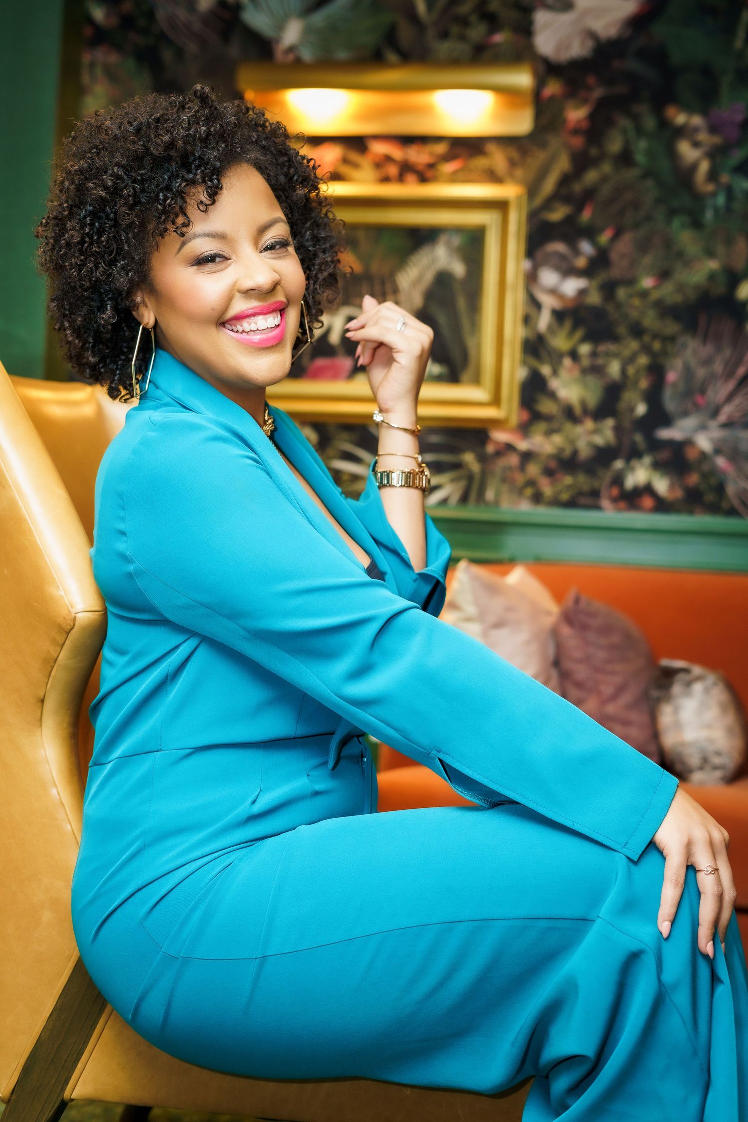 Elegant woman in teal suit sits smiling in gold chair against vibrant floral background.