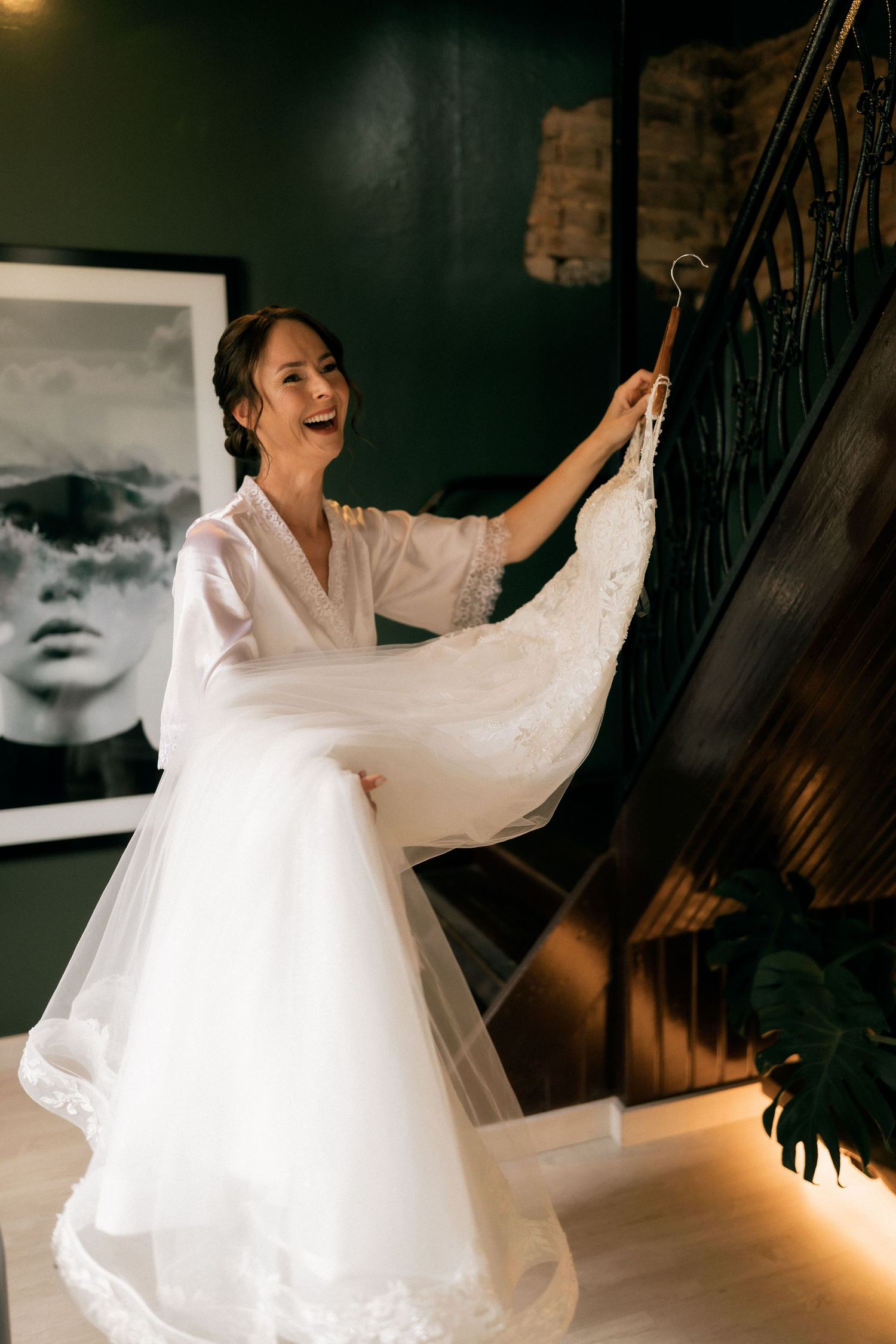 Joyful bride in white robe holding lace wedding dress on hanger at bottom of dark staircase.