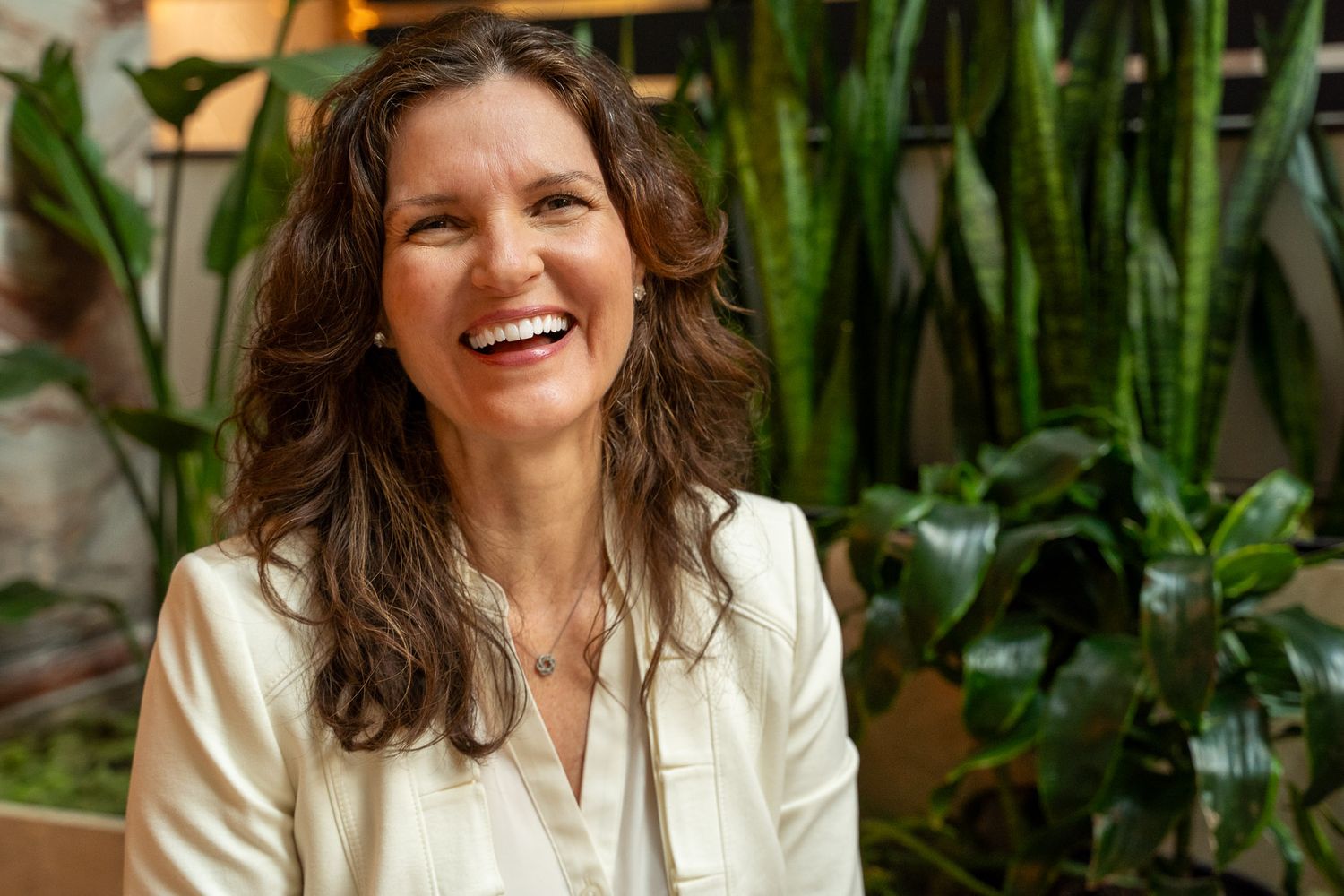 Woman with long brown hair laughing joyfully, wearing a white blazer, surrounded by lush green tropical plants.