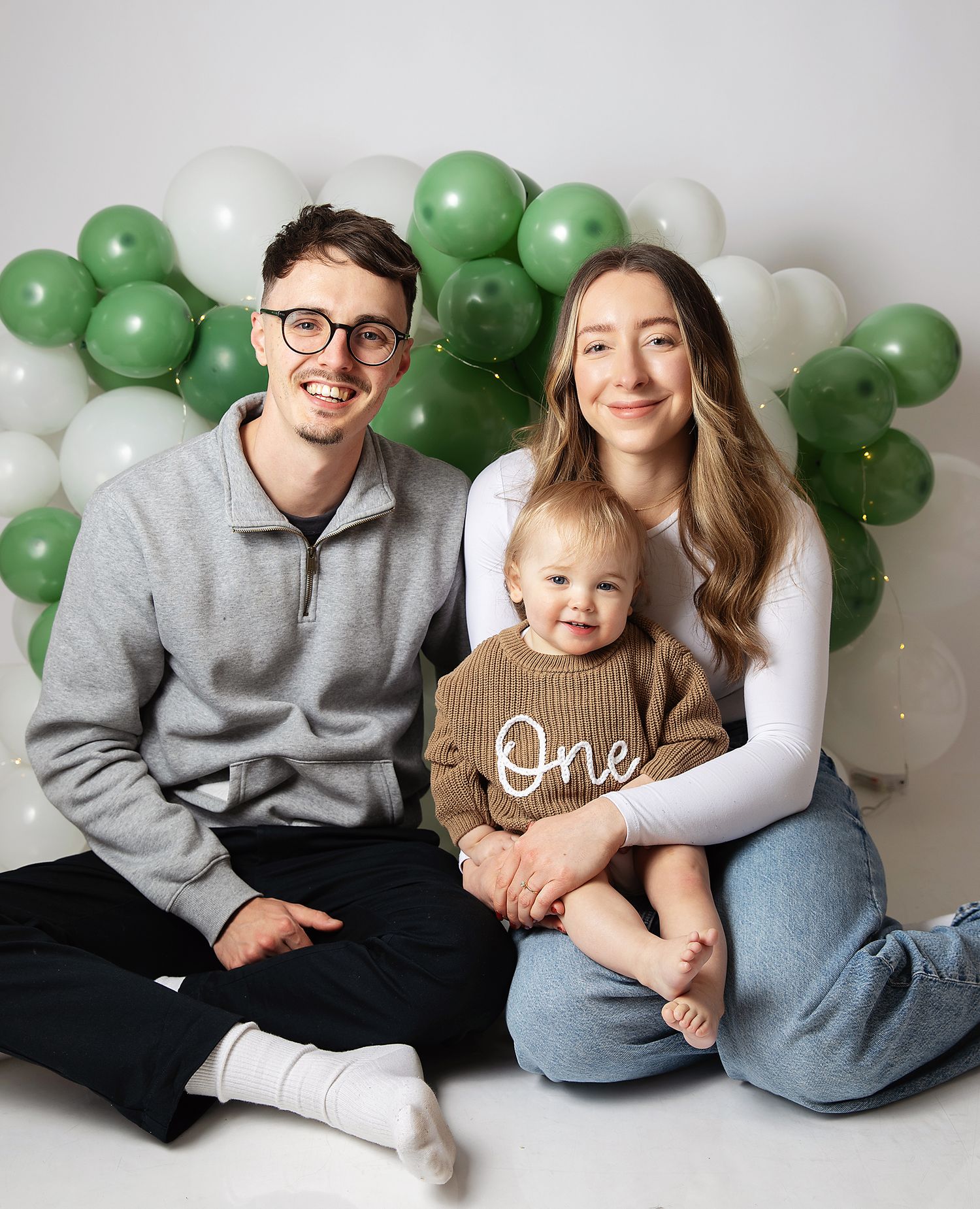 Family photo with parents and baby celebrating first birthday with green and white balloon backdrop.