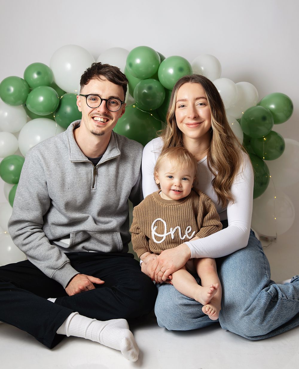 Family photo with parents and baby celebrating first birthday with green and white balloon backdrop.