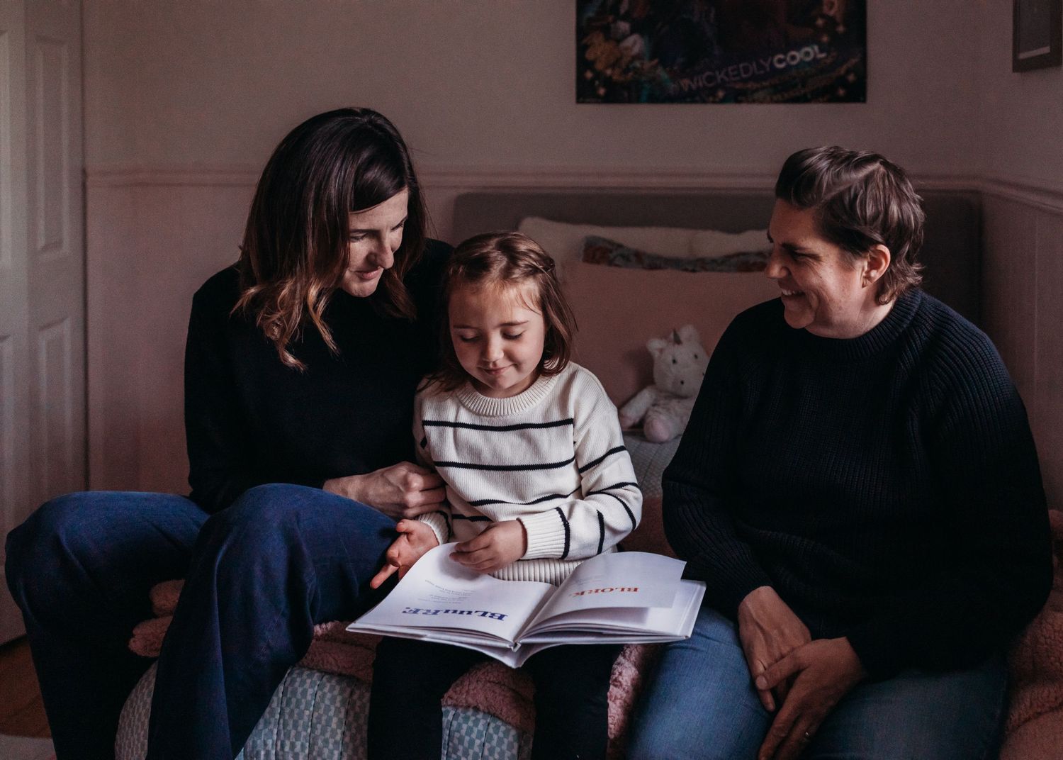Two women and a young girl sit together on a bed, smiling while reading a book together indoors.