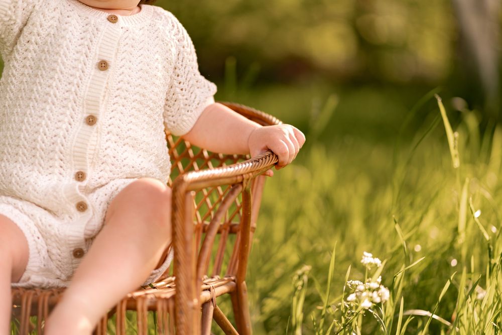 Baby in knit romper sitting on wicker chair in sunny green grass outdoor setting during a childhood photography session in Lincolnshire.