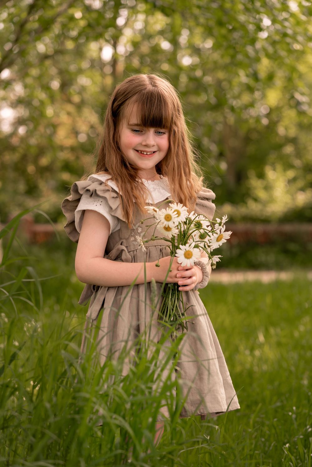 Young girl in vintage dress holding white daisies bouquet, smiling outdoors during a childhood photography session in Lincolnshire.